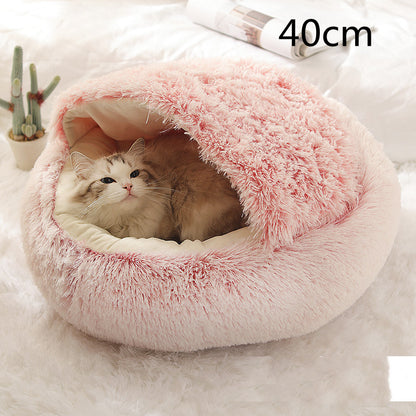 Pink plush round pet bed with the hood raised over a cat resting on the cream lining, a small cactus plant behind, the option size printed in the corner