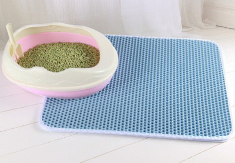 Light blue honeycomb litter mat with a white edge on pale floorboards, beside a pink and cream litter tray holding a scoop and green litter