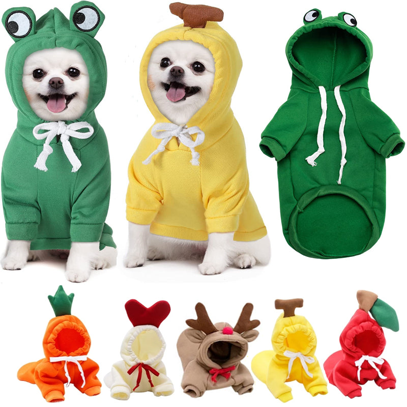 Two small white dogs in green frog and yellow banana hoodies, an unworn green frog hoodie photographed from the back showing its hood, two sleeves and drawstrings, and a row of five more unworn hoodies below: carrot, cream with a padded red heart, tan with antlers, banana and apple